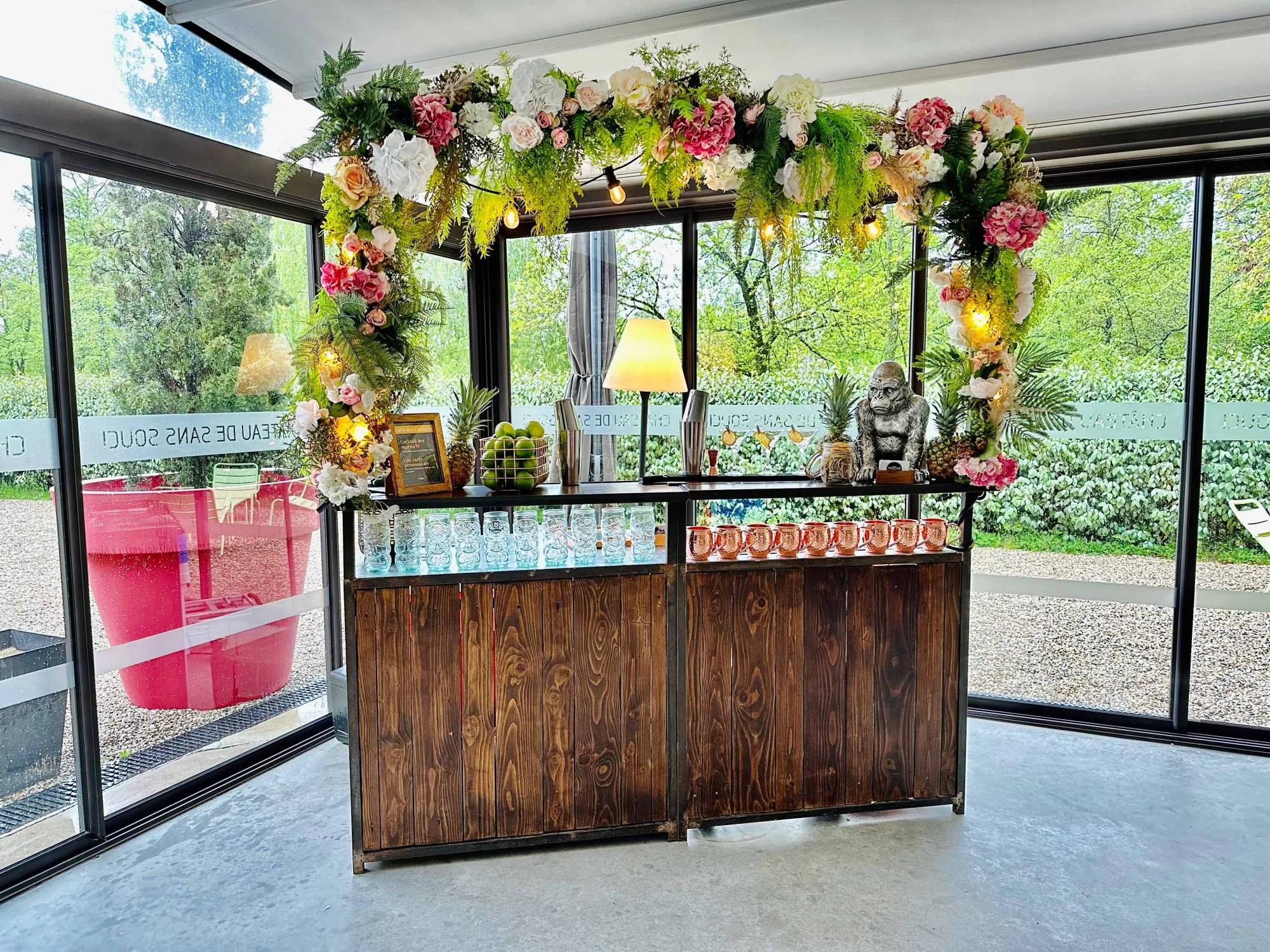 Bar à cocktails en bois sous une arche de fleurs, avec des verres et des tasses en cuivre alignés