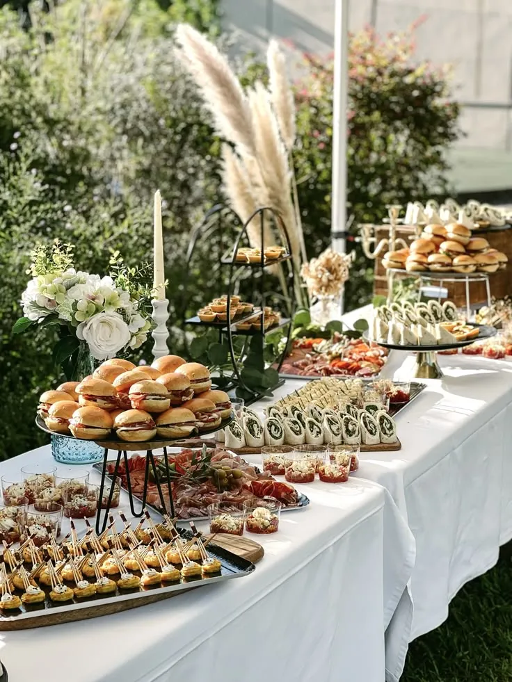 Buffet dressé en extérieur sur nappe blanche, avec mini-burgers, verrines, brochettes et bouquets de fleurs